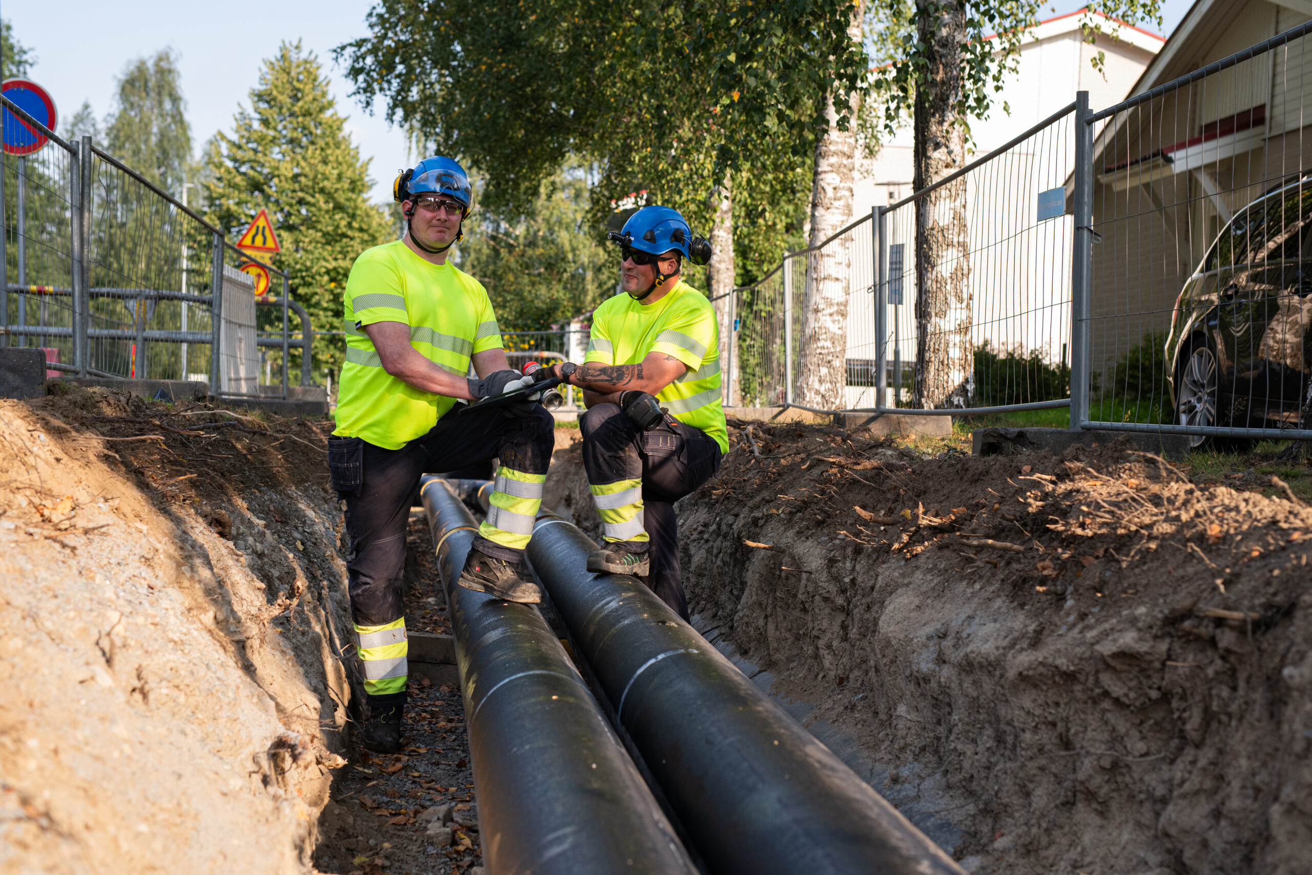 Miehet katsovat kameraa kaukolämpöputken luona.
